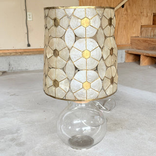 1970s capiz shell and brass drum lampshade in a hexagon flower pattern, sitting on a clear round glass lamp base, shown unlit on a concrete floor