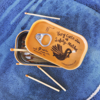 Orange Very Cute Tin of Safety Matches by Tinned Candle, lid lifted to show orange-tipped matches inside, fan-tailed bird illustration on the lid, loose matches on a blue ceramic plate