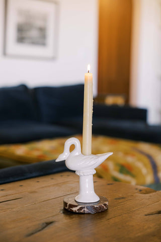 Cream ceramic swan candlestick holder with a lit ivory taper, set on a brown-and-white marble coaster on a worn wood coffee table in front of a navy sofa