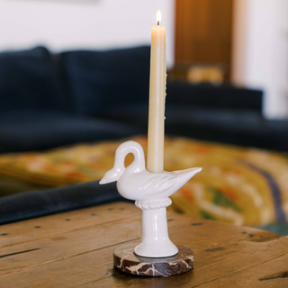 Cream ceramic swan candlestick holder with a lit ivory taper, set on a brown-and-white marble coaster on a worn wood coffee table in front of a navy sofa
