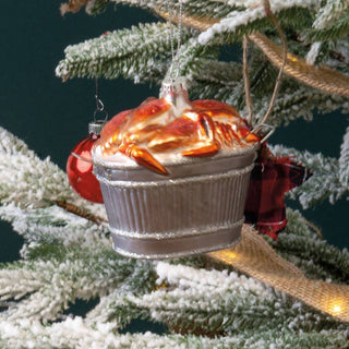 The glass crab bushel ornament hanging on a frosted Christmas tree branch beside a red ball ornament and a plaid bow