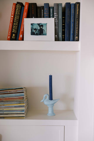 Light blue ceramic swan candlestick holder with an unlit navy taper on a white built-in shelf below a row of books