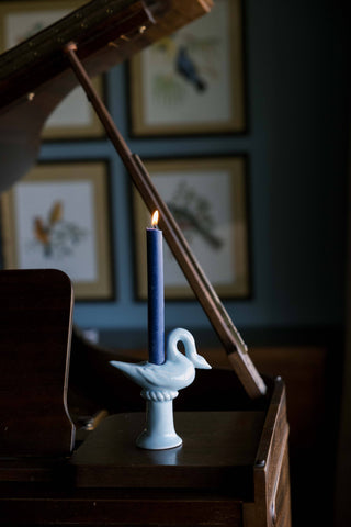 Light blue ceramic swan candlestick holder holding a lit navy taper candle on a dark wood piano, framed bird prints on a blue wall behind