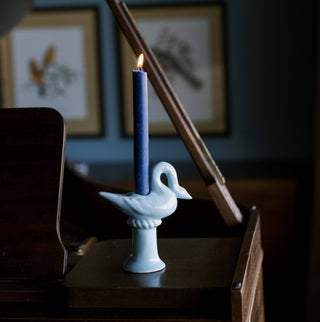 Light blue ceramic swan candlestick holder holding a lit navy taper candle on a dark wood piano, framed bird prints on a blue wall behind
