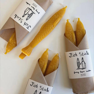 Fish Sticks beeswax tapers in kraft-paper wraps with illustrated 'Fish Sticks fancy taper candles' labels, one unwrapped taper lying flat to show its scales, fins and tail
