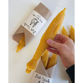 A hand holding one Fish Sticks beeswax taper to show its full length — sculpted scales, fins and a fanned tail — with wrapped pairs behind it