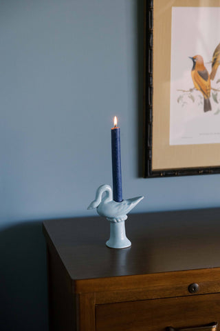 Light blue ceramic swan candlestick holder with a lit navy taper on a walnut dresser beneath a framed bird print on a blue wall