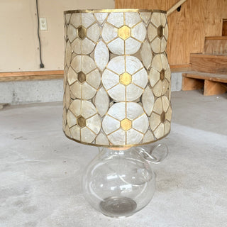 1970s capiz shell and brass drum lampshade in a hexagon flower pattern, sitting on a clear round glass lamp base, shown unlit on a concrete floor