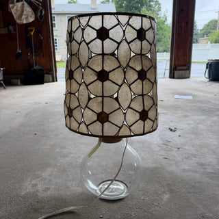 1970s capiz shell and brass drum lampshade on a clear round glass base, backlit in an open garage doorway so the shell panels glow translucent