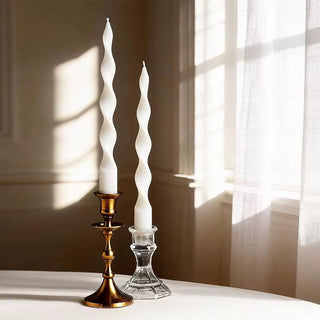 Pair of ivory twisted spiral taper candles in brass and glass holders on a sunlit table.