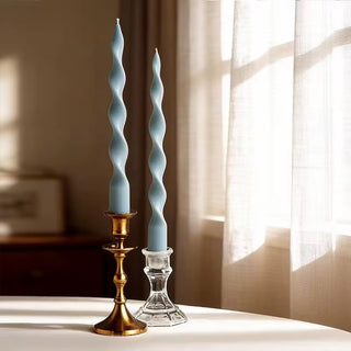 Pair of smoke blue twisted spiral taper candles in brass and glass holders by a sunlit window.