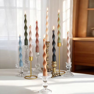 Twisted spiral taper candles in five colors, several lit, clustered in brass and glass holders on a sunlit table.
