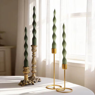 Two olive green twisted spiral taper candles in ornate gold candlesticks on a white table by a window.