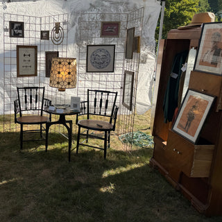 Wren Supply Co. booth sitting vignette — the lit 1970s capiz and brass lamp on a small black table between a pair of antique black cane-seat chairs, grid walls of framed art behind (Cretan figure studies, swan, embroidered pup, brass wall calendar, wood frames), and a wardrobe with a straw garden hat on top