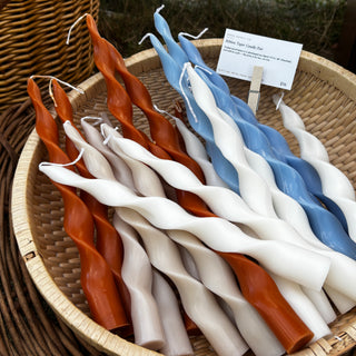 Twisted ribbon taper candles in caramel, milk tea, ivory, and smoke blue piled in a woven basket with a small price card clipped to the edge — Wren Supply Co. booth