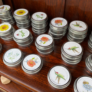 Stacks of Garden Sprinkles seed tins on a wooden shelf — painted lids showing sunflower, good luck clover, wildflowers poppy, hummingbird, forget-me-not, butterfly, and honeybee — Wren Supply Co. booth