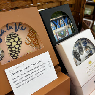 Three Idlewild Co. porcelain trinket dishes in their window boxes propped in a picnic basket — Sea La Vie with painted shells, Sardines with blue fish, and Oysters — with a museum-style price card in front, Wren Supply Co. booth