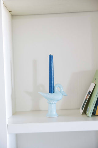 Side view of the light blue ceramic swan candlestick holder holding an unlit blue taper on a white shelf, the swan facing right