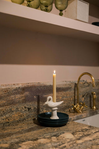 Cream ceramic swan candlestick holder with a lit ivory taper on a stack of dark green plates on a granite countertop beside a brass faucet