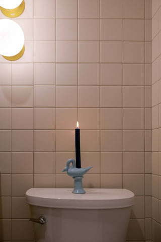 Light blue ceramic swan candlestick holder with a lit dark taper on the back of a toilet against pale pink square tile, a globe wall light above