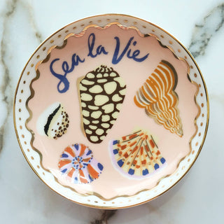 Sea La Vie blush porcelain trinket dish hand-painted with assorted seashells and cobalt 'Sea la Vie' lettering, gold-lined scalloped rim, shown from above on marble