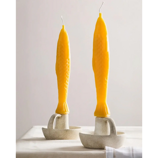 Two Fish Sticks beeswax taper candles standing tail-down in textured white chamberstick holders on a linen-covered table