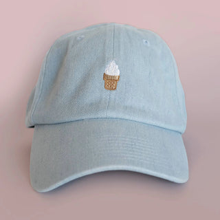 Light denim ice cream cone baseball hat styled on a soft pink background, front view of the embroidered soft serve cone