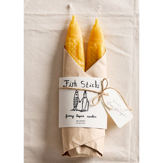 A pair of Fish Sticks beeswax taper candles by Tinned Candle, fish heads poking out of a kraft-paper wrap with an illustrated 'Fish Sticks fancy taper candles' label, tied with twine and a gift tag, lying on natural linen