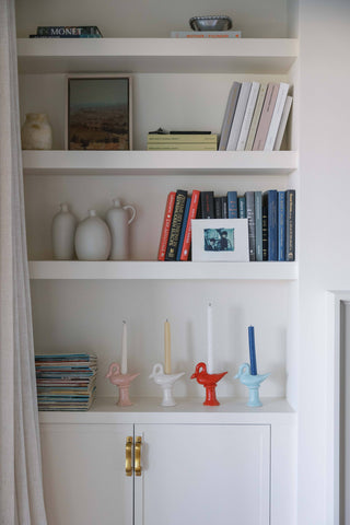 Four St. Bingo ceramic swan candlestick holders in a row on a white shelf, in pink, cream, red and light blue, each holding a taper; Wren carries the blue and cream