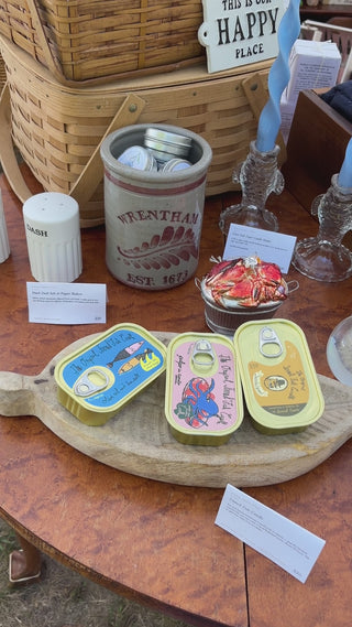Two smoke blue twisted ribbon taper candles standing in clear glass fish taper candle holders on a wooden table at the Wren Supply Co. booth, tinned fish candles in front