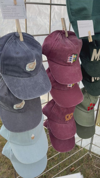 Six embroidered baseball hats clipped to a grid wall in three color columns — navy and light blue, burgundy, forest and light green — Wren Supply Co.