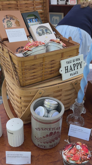 Slow pan up to the glass crab bushel ornament — a heap of red glass crabs in a silver bushel basket — then a hand lifts it, turns it slightly, and sets it back down at the Wren Supply Co. booth