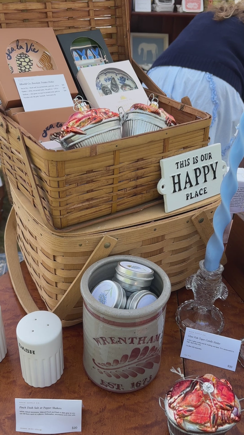 Slow pan up to the glass crab bushel ornament — a heap of red glass crabs in a silver bushel basket — then a hand lifts it, turns it slightly, and sets it back down at the Wren Supply Co. booth