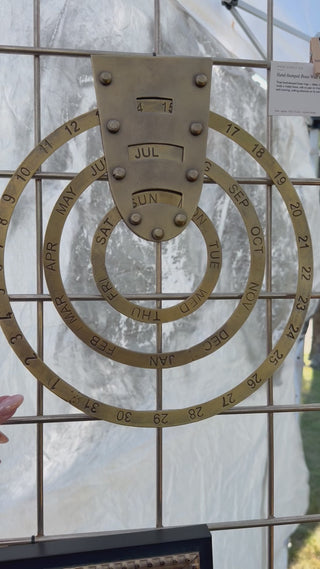 The hand-stamped brass wall calendar's three rings being spun — outer ring for the month, middle ring for the date, inner ring for the day of the week