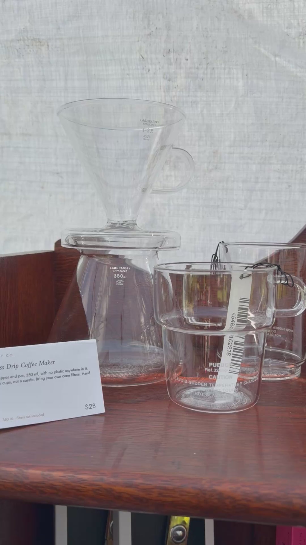 Puebco glass drip coffee maker — cone dripper on its 350 ml pot — beside stacked borosilicate glass mugs and glass salt and pepper shakers on a wooden shelf at the Wren Supply Co. booth